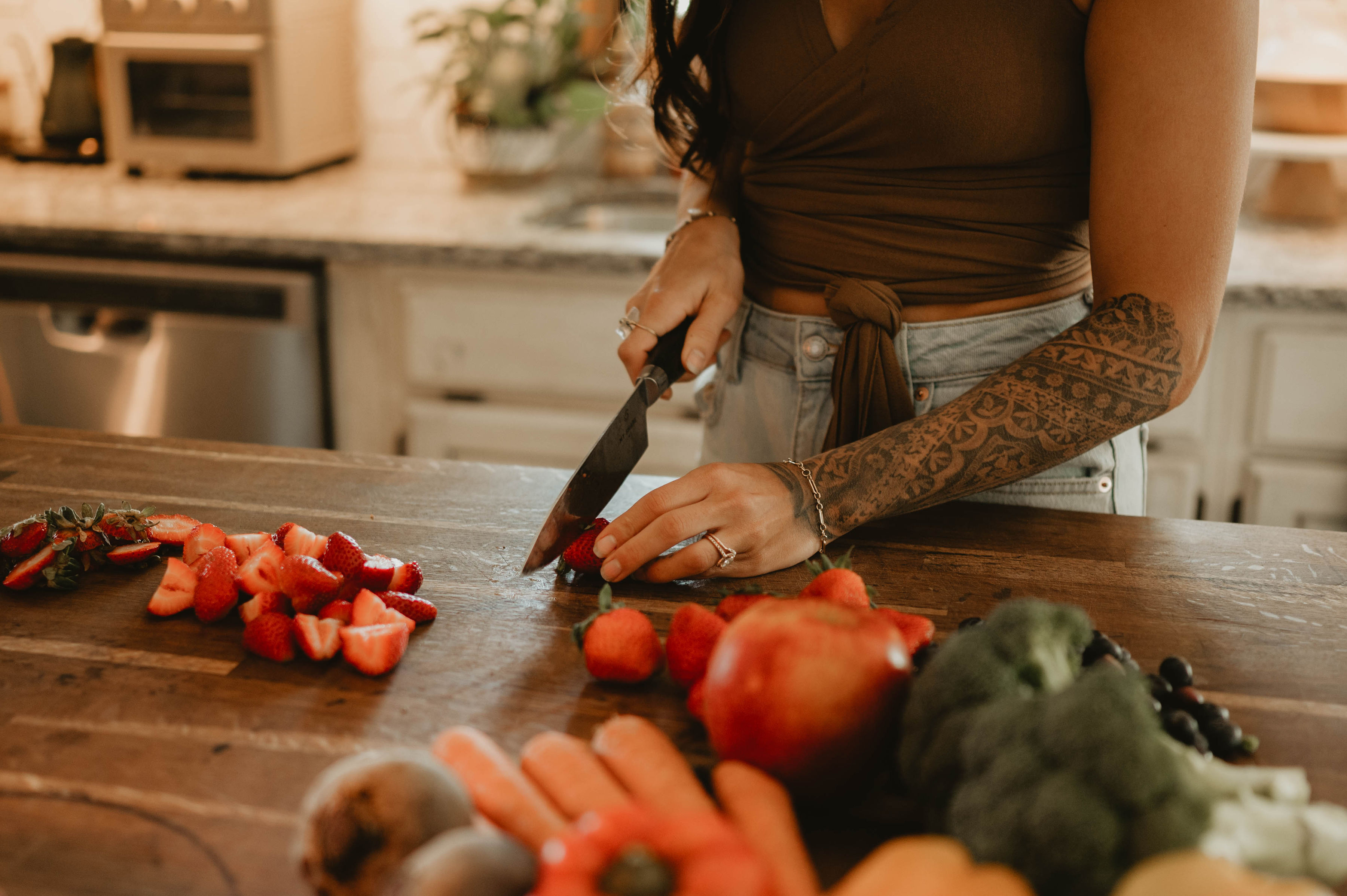 A woman with tattoos and a brown crop top stands at a kitchen island, cutting strawberries with a large knife.