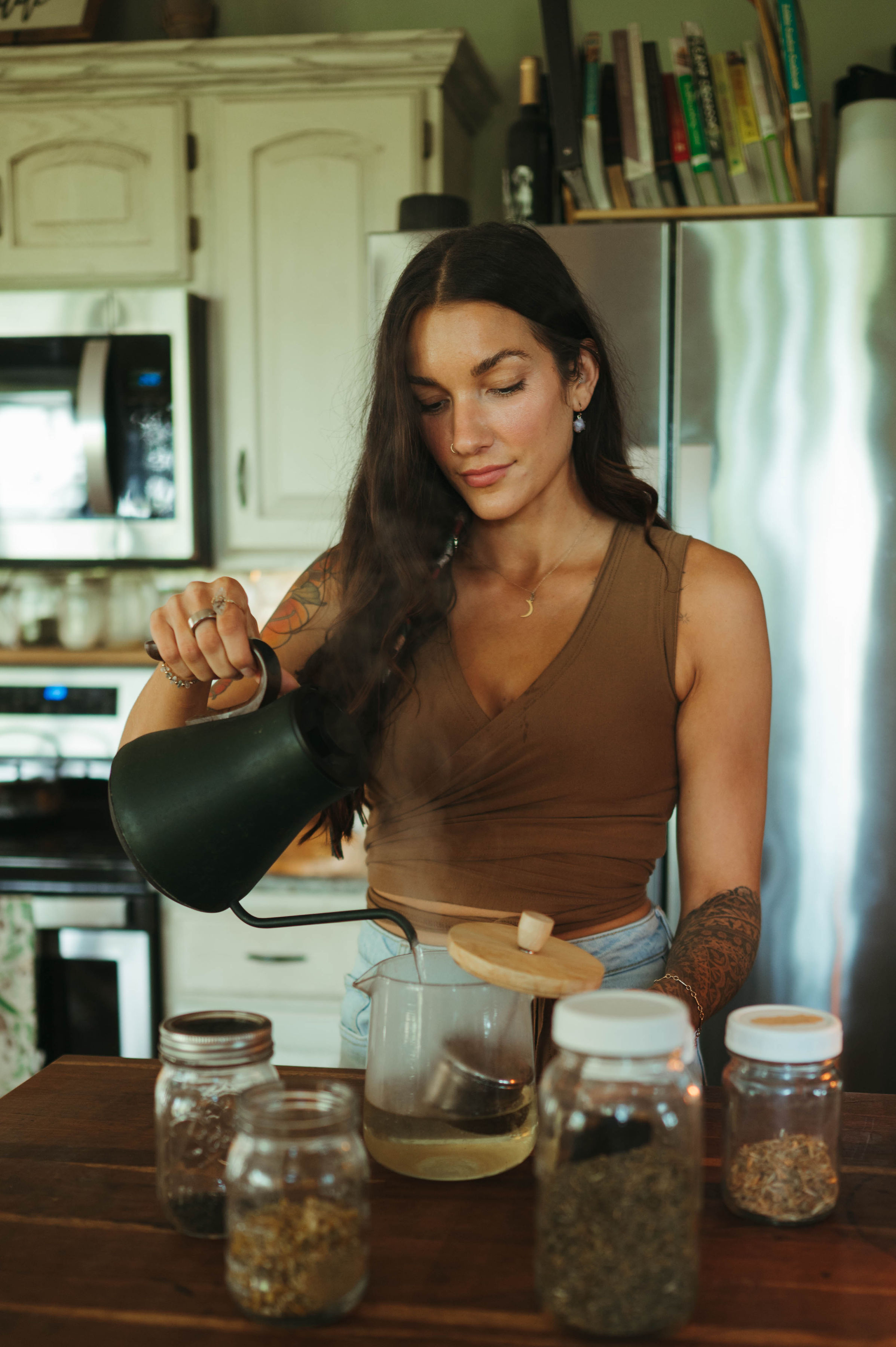 A woman with long dark hair pours water from a black kettle into a glass teapot on a kitchen counter.